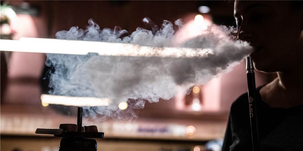 Zoll stellt in Kiosk in Recklinghausen ein Kilo Betäubungsmittel sicher Eine junge Frau im Schatten atmet sichtbar den Rauch einer Shisha aus.