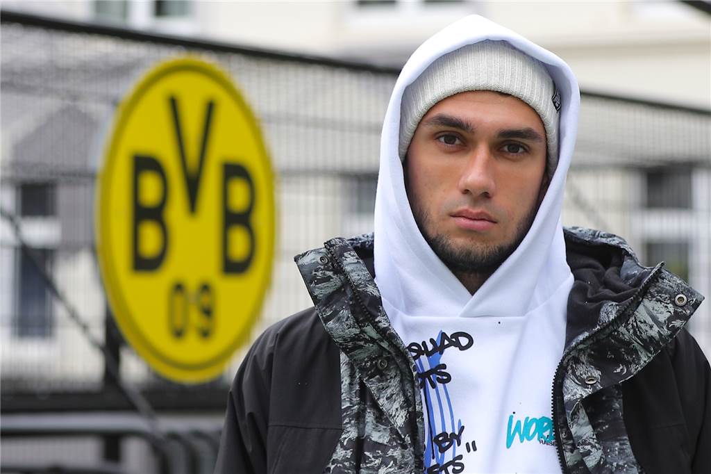 Vom Kriegsflüchtling zum Profifußballer: Alan Aussi kann dank BVB seinen Traum leben Der Ukrainer Alan Aussi steht vor einem BVB-Wappen und schaut in die Kamera.