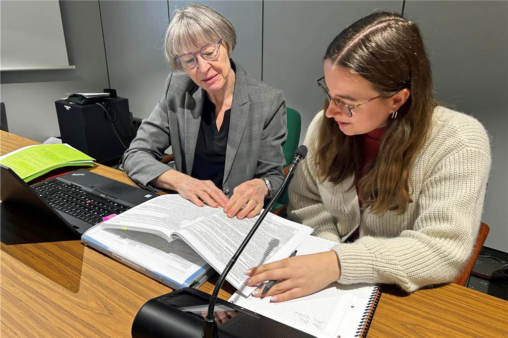 Manuela Gerdemann (rechts) und Lara Overmann sitzen an einem Tisch, auf dem vor ihnen ein Laptop und ein Mirofon stehen, und scheuen in Unterlagen.