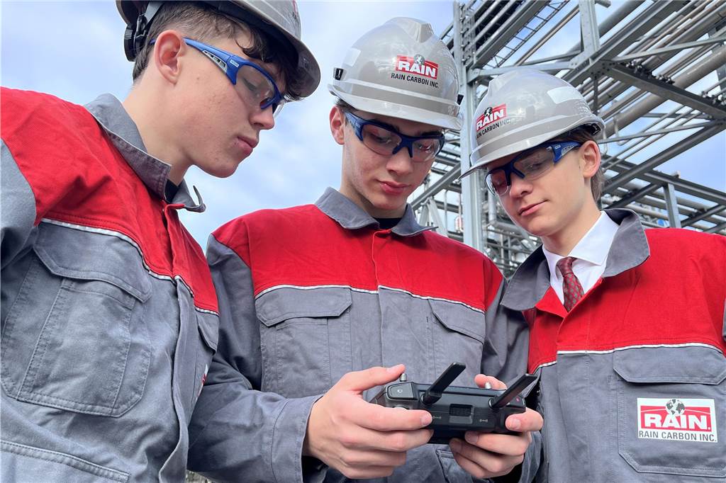 Max Hayden, Damien Rachuba und Julius Janke (v.l.) schauen auf das Display einer Fernbedienung. Sie tragen Arbeitsanzüge in Grau-Rot und graue Helme.
