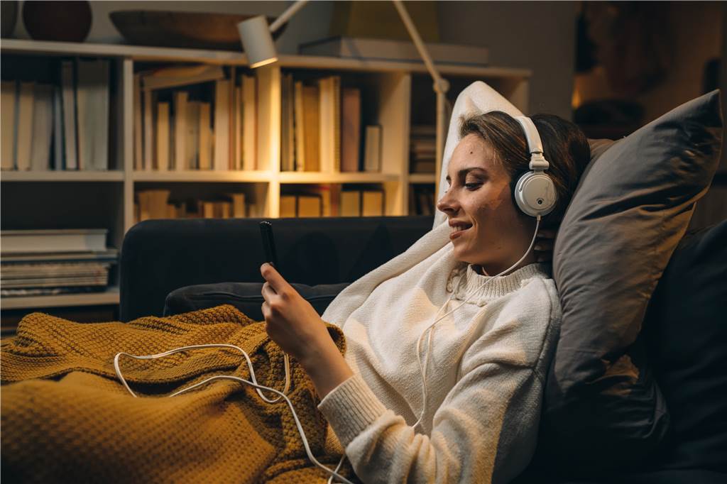 Die dunkle Jahreszeit perfekt nutzen: Freizeitaktivitäten im Dezember frau liegt gemütlich unter einer decke auf der couch und hat kopfhörer auf