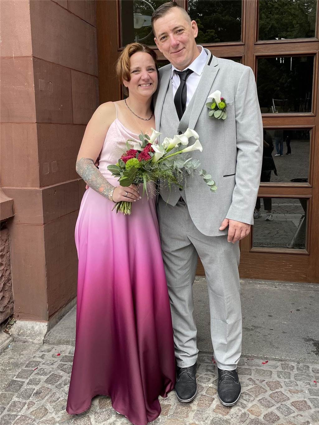 Freudentränen bei den Kindern: Stefan und Manuela sind verheiratet Michaela präsentiert ihr Brautkleid. Der Stoff zeigt einen Farbverlauf von Rosa nach Pink. Ihr Bräutigam Stefan steht neben ihr.