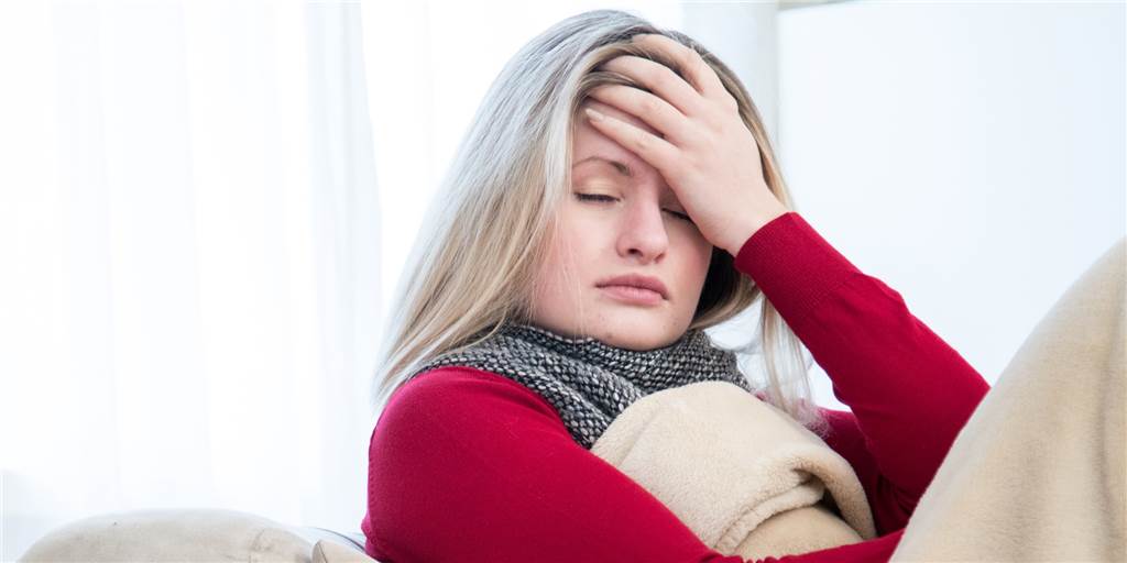 Nur Zufall? Warum manche Menschen kein Corona hatten Eine Frau liegt unter einer Decke eingekuschelt auf der Couch, hat die Augen zu und hält sich die Hand an den Kopf.