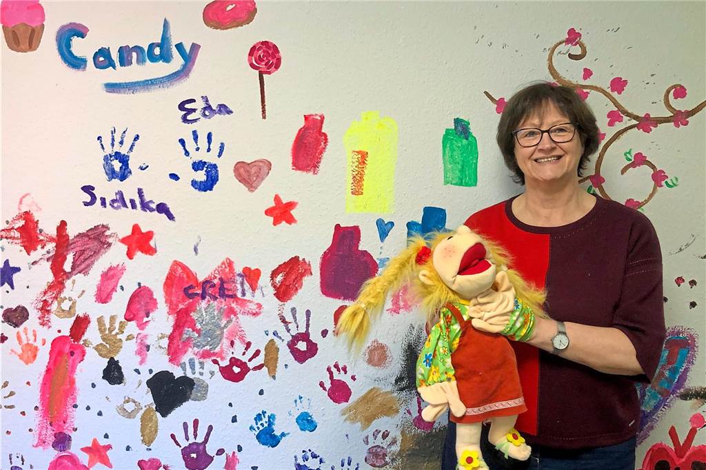 Anna Pein steht vor einer Wand mit bunten Handabdrücken und hält eine Handpuppe im Arm.