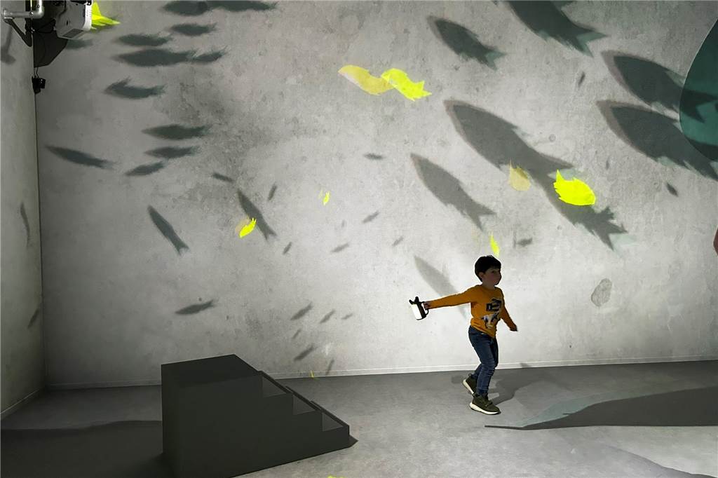 Was können Kinder in der neuen Familien-Ausstellung im U erleben? Ein Junge mit einer Lampe in der Hand in einem grauen Raum, über dessen Wände "Schattenfische" schwimmen.