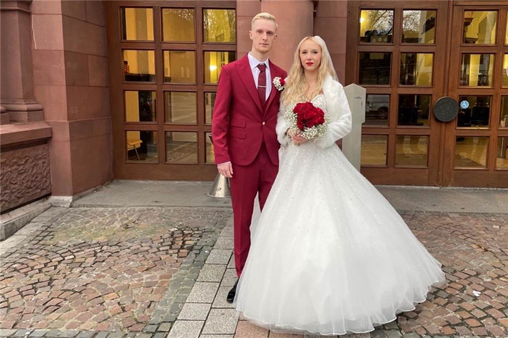 Ann-Cathrines Kleid hat einen breiten Tüllrock in Weiß. Sie steht neben Christopher vor dem Alten Stadthaus. Der Bräutigam trägt einen dunkelroten Anzug. In Ann-Cathrines Brautstrauß sind Rosen - ebenfalls in Rot.