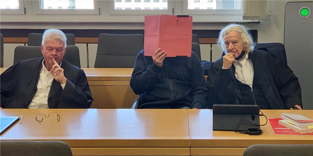 Mütter zum Missbrauch gedrängt: Ex-Tennislehrer wieder vor Gericht Die Verteidiger Wolfgang Zeitler und Siegmund Benecken neben dem Angeklagten im Essener Landgericht, der sich einen roten Aktendeckel vors Gesicht hält.