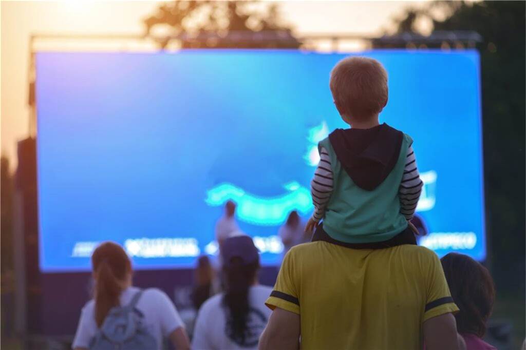 Filmgenuss unterm Sternenhimmel: 9 Open Air-Kinos in NRW kind sitzt auf den schultern eines erwachsenen und guckt auf eine leinwand