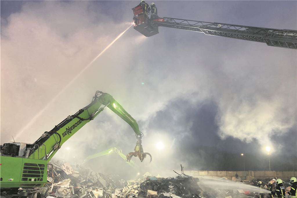 Feuerwehrleute stehen an einem Schrotthaufen und richten Löschschaum darauf. Ein Bagger hebt Teile des Haufens zur Seite.