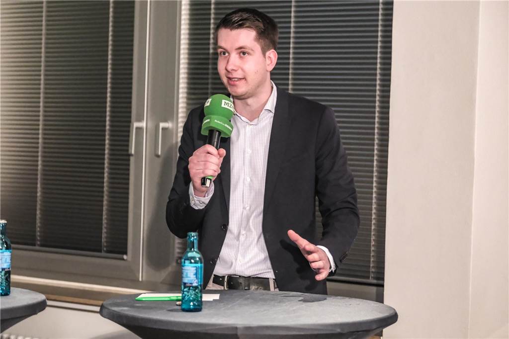 Tom Roehl steht an einem grauen Stehtisch und spricht in ein grünes Mikrofon. Hinter ihm ist eine Fensterzeile zu sehen.