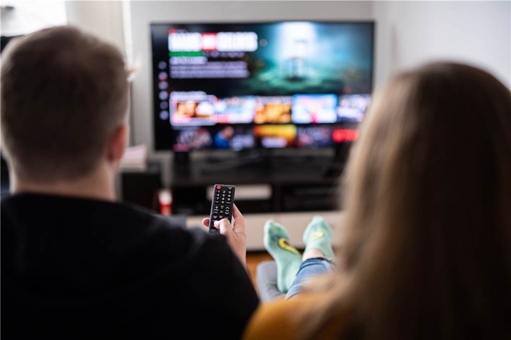 Die zehn größten Stromfresser im Haushalt – und wie man ihren Verbrauch drosselt Ein Paar sitzt vor dem Fernseher, der Mann hält die Fernbedienung.
