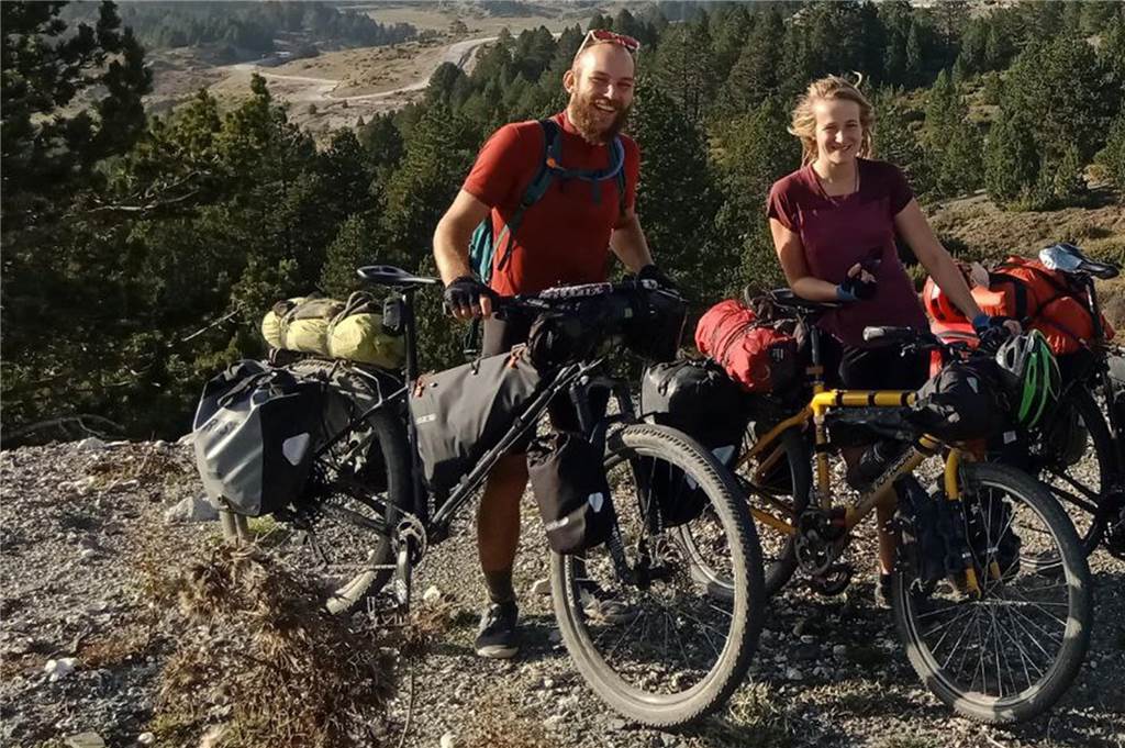 Pia und Veit stehen mit ihren Fahrrädern auf einem Hügel. Hinter ihnen eine Berglandschaft. Beide halten ihre Fahrräder bei sich. 