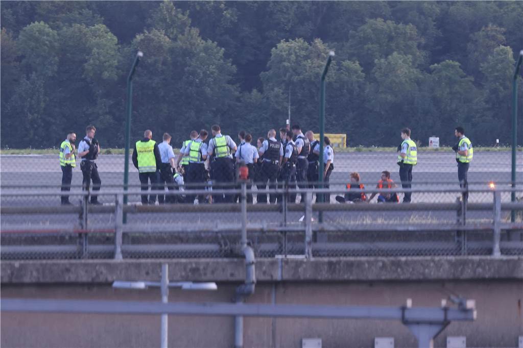 Mitglieder der Letzten Generation blockieren Düsseldorfer Flughafen: Störungen im Flugverkehr Polizisten stehen auf dem Flugfeld und versuchen Aktivisten der Gruppe Letzte Generation vom Asphalt zu lösen, nachdem sie sich festgeklebt haben.