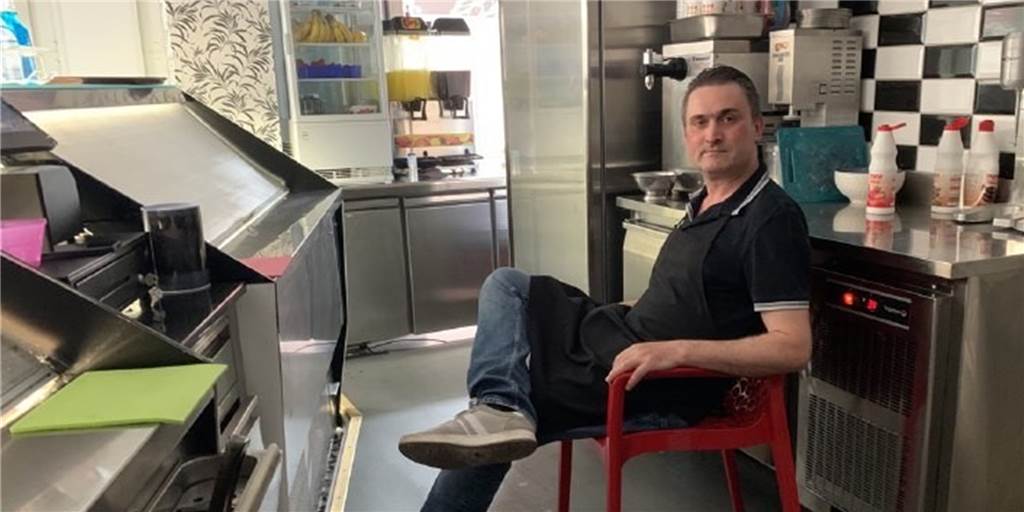 Nach Einbruch in Eisdiele: Fadil Faslija schläft auf Plastikstuhl in seinem Laden Ein Eisverkäufer sitzt auf einem roten Plastikstuhl hinter der Theke eines Eiscafés. Rechts hinter ihm ist eine Theke, auf der verschiedene Soßen stehen, links vor ihm die Rückseite der Eistheke.