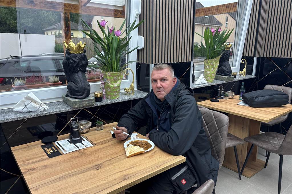 Silvan Fischbach sitzt an einem Tisch im frisch sanierten Grillhaus Hades. Vor ihm steht ein Teller mit Brot und Fleisch. In der Hand hält er eine Gabel.