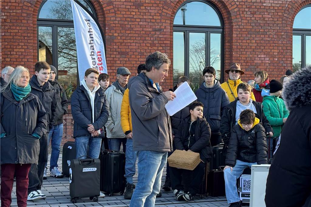 Ein älterer Herr hält eine Rede vor dem Bahnhof. Er liest einen Text vor. Im Hintergrund sind weitere Teilnehmer vor dem Bahnhof zusehen.