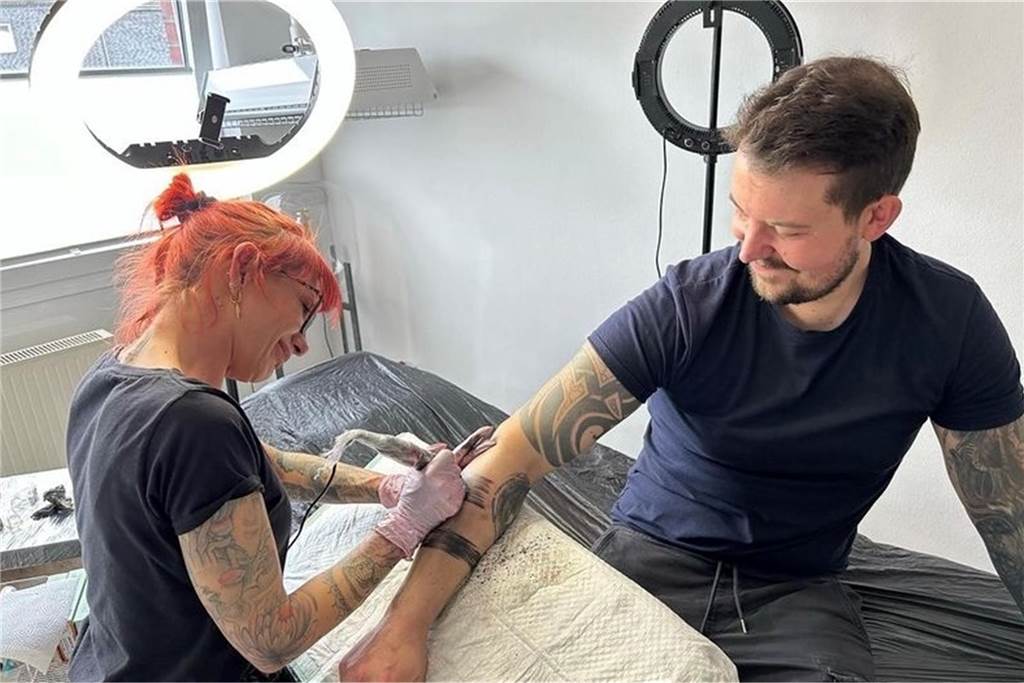 Eine Frau sticht einem Mann ein Tattoo auf den rechten Unterarm.