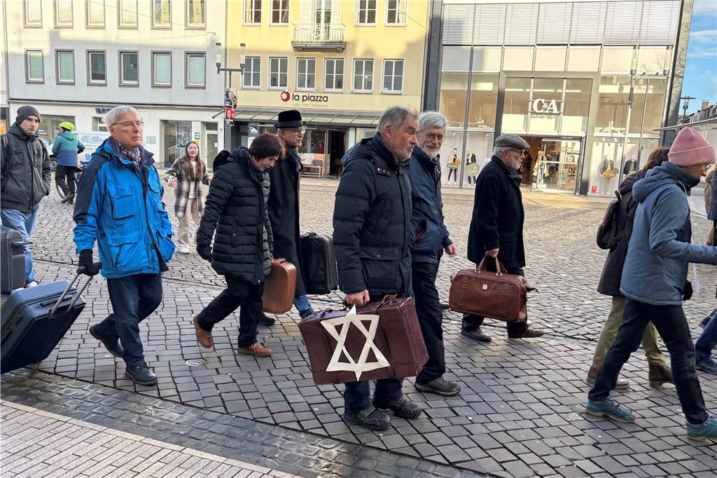 Personen aus der Mitte des Marsches laufen über den Marktplatz mit Koffern in der Hand. Einer der Koffer trägt einen jüdischen Stern. 