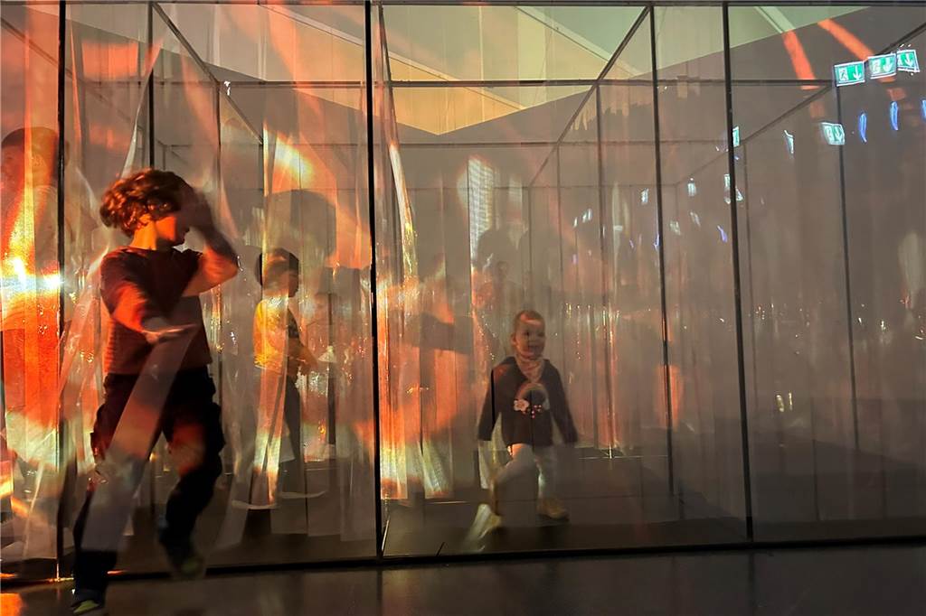 Was können Kinder in der neuen Familien-Ausstellung im U erleben? Drei Kinder huschen durch einen Raum, in dem durchsichtige Stoffbahnen hängen.