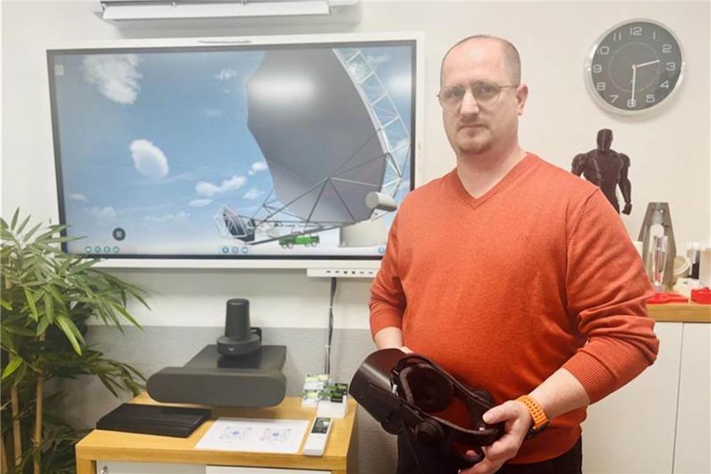 Tobias Will hält eine VR-Brille in der Hand ist und ist im Vordergrund zu sehen. Hinter ihm ein Fernseher, mit dem Bild, welches man durch die Brille sieht.