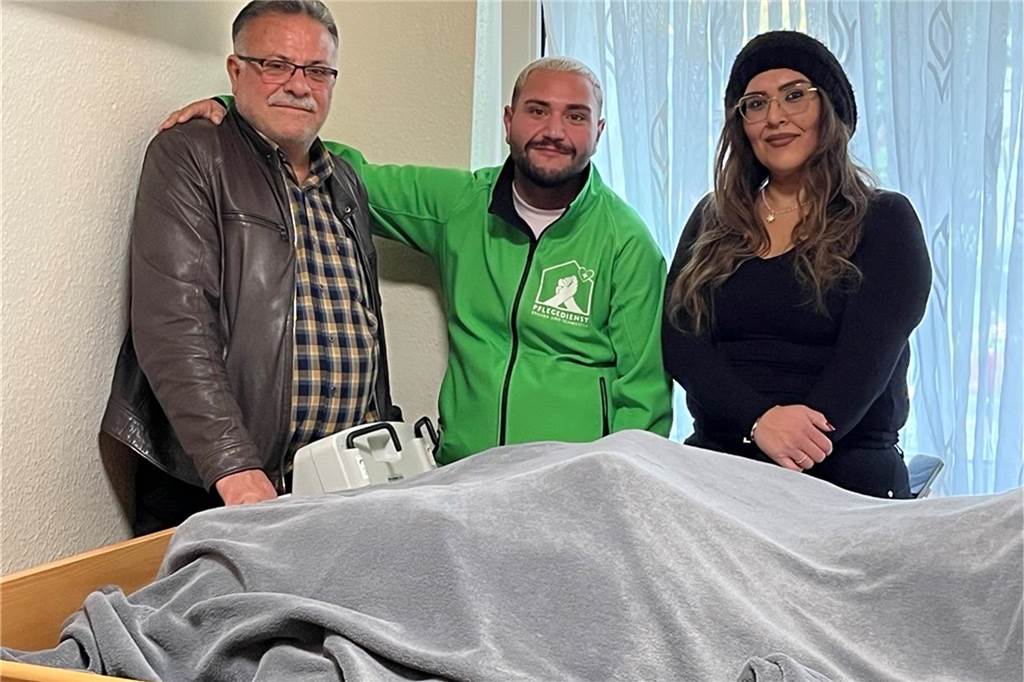 Vater und Sohn mit Pflegerin Tuba, die auch zur erweiterten Familie gehört, stehen am Bett der Patientin Frau Kaya. Sie legen viel Wert auf einen familiären Umgang.