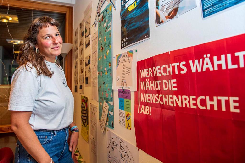 Katharina Friedrich steht vor einer Wand, an der verschiedene Plakate hängen. Im Vordergrund hängt ein rotes Plakat, auf dem in weißen Großbuchstaben steht: "Wer Rechts wählt, wählt die Menschenrechte ab!".