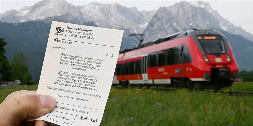 Tarifwirrwarr um 9-Euro-Ticket: Wo Reisende genau hinschauen müssen Ein Mann hält ein 9-Euro-Ticket in der Hand, im Hintergrund fährt ein Zug der Deutschen Bahn vor der Bergkulisse des Wettersteingebirges vorbei (gestelltes Foto).