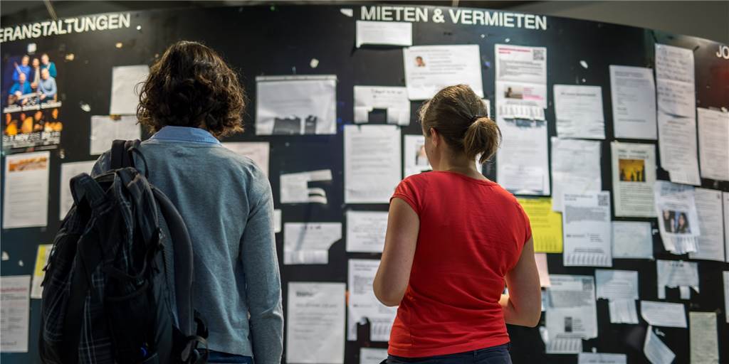 Zwei Studentinnen betrachten die Wohnungsanzeigen am Schwarzen Brett einer Universität.