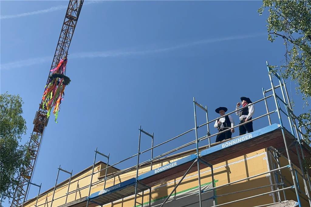 OGS feiert Richtfest: Acht-Millionen-Projekt soll im Herbst fertig sein Zwei Männer in traditioneller Tracht stehen auf einem Gerüst vor einem gelben Gebäude. Im Hintergrund ist ein Kran mit einem bunten Kranz zu sehen.